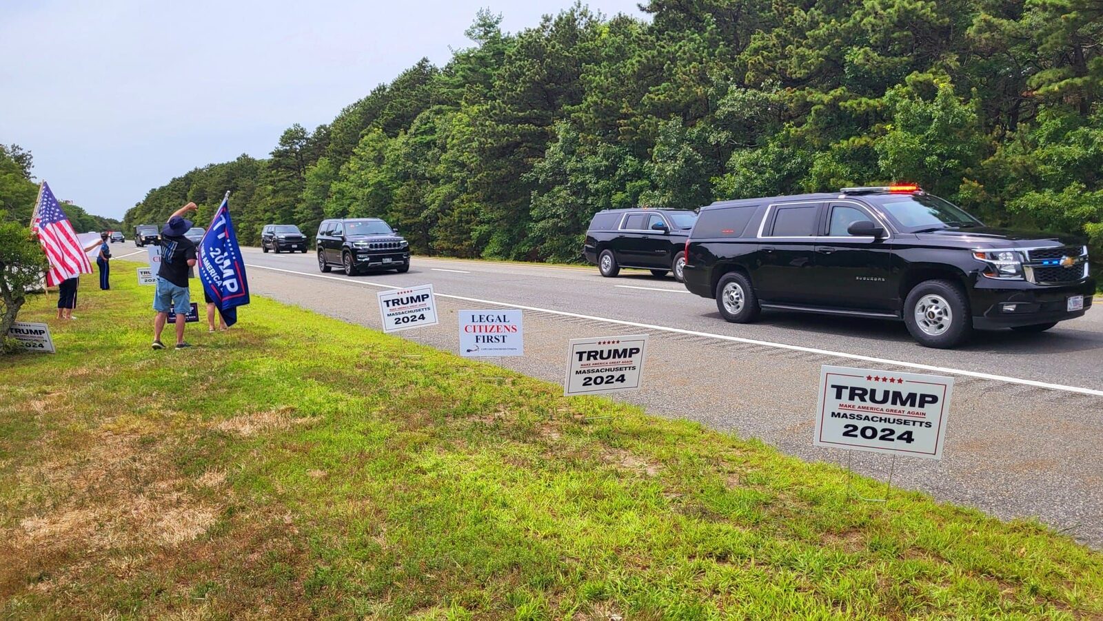 Roadside stand-out — flags up and “Legal Citizens First” signs in the ground as the motorcade passes.